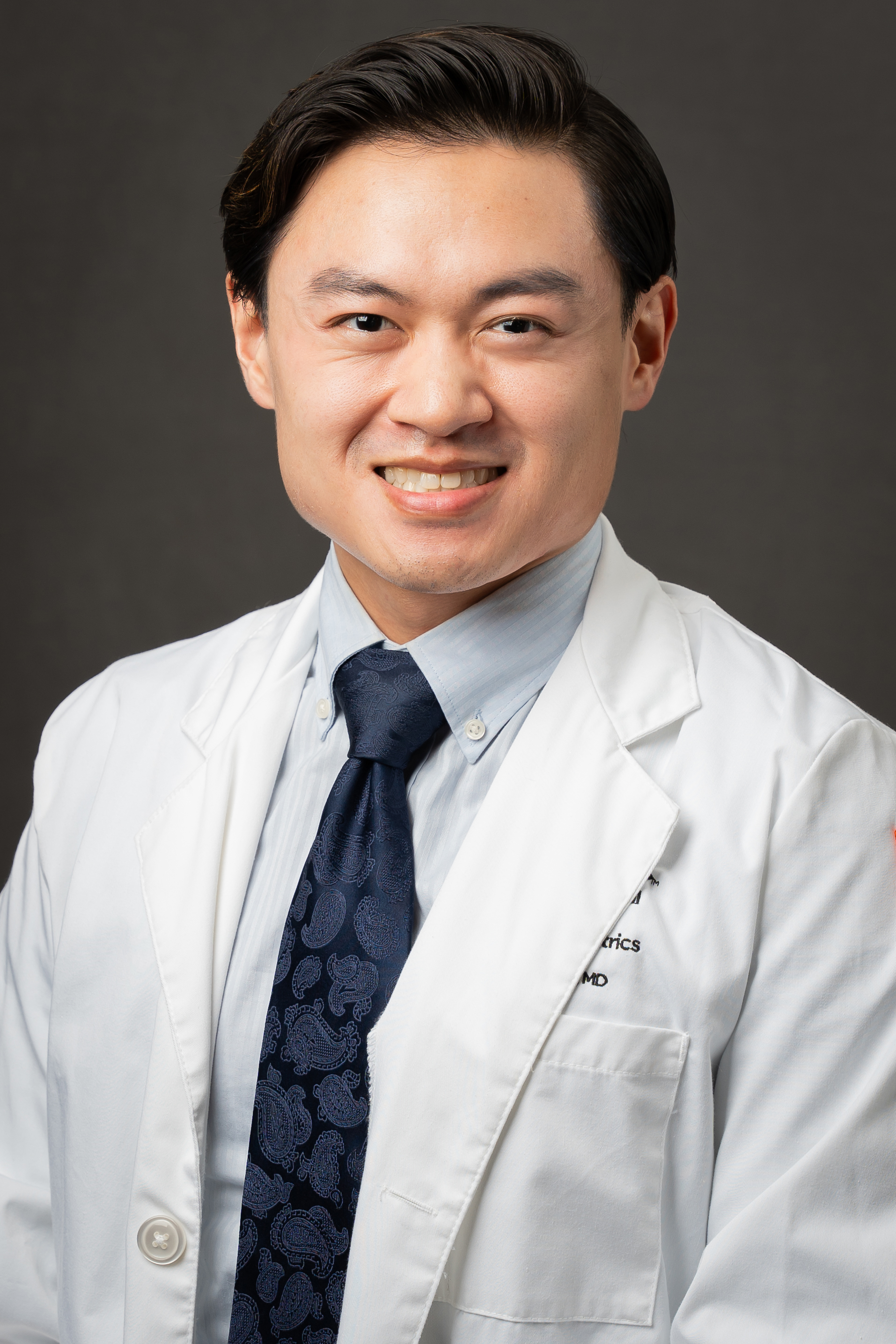 kevin kuo is seated wearing a tie, blue shirt, and white lab coat while smiling for the camera.